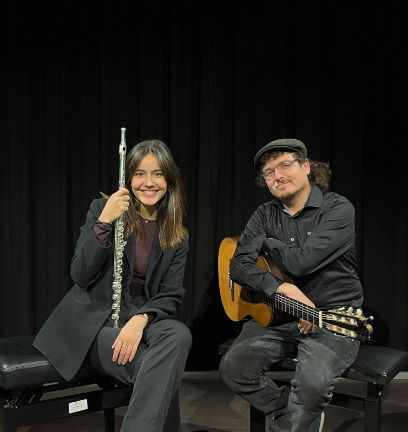 Vor schwarzem Hintergrund sitzen zwei Musiker*innen, beide schwarz und elegant gekleidet. Die Person links hat lange braune Haare und eine Querflöte in der Hand. Die Person rechts trägt einen Hut und hat eine Gitarre auf dem Schoss.