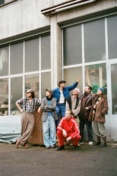 Vor einem Fabrikgebäude mit hohen Fenstern stehen sieben junge Menschen. Einer davon deutet nach rechts oben, alle anderen schauen nach links oben.