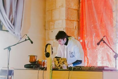 Vor einer Backstein-Wand mit bunten Vorhängen steht ein junger Mann mit weissem Pullover vor einem Tisch mit diversen Geräten wie DJ-Pult, Laptop, Klangkörpern, Kopfhörern und zwei Mikrofons. 