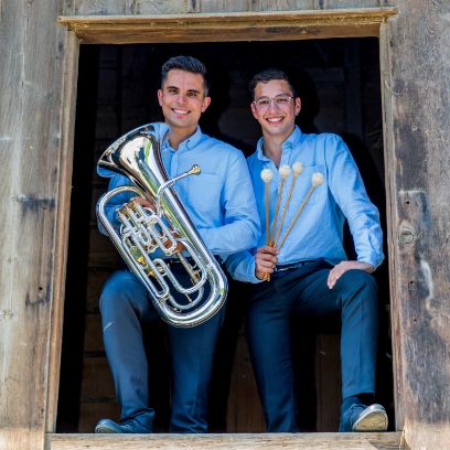 Zwei junge Männer stehen in einer grossen Holzluke und schauen herunter. Beide tragen blaue Hosen und hellblaue Hemden. Der Mann links hält ein Euphonium in den Händen, der Mann rechts hält vier Schlägel in der Hand. Beide lachen in die Kamera.
