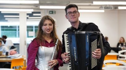 Zwei Personen vor dem Hintergrund einer Universitätsmensa. Die linke Person ist eine Frau mit einer Querflöte in der Hand, die rechte Person ein Mann mit Brille und einem Akkordeon in der Hand.