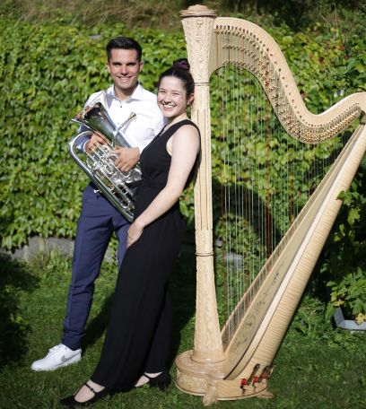 Vor einer grünen Hecke stehen zwei Personen. Vorne steht eine Frau an eine Harfe angelehnt. Sie trägt ein schwarzes Kleid. Hinten steht ein Mann mit weissem Hemd, der ein Euphonium in der Hand hält. Beide lächeln in die Kamera. 