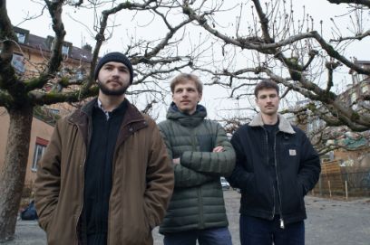 Auf einem Kiesplatz mit Bäumen und dahinter einigen Wohnhäusern im Hintergrund stehen drei junge Männer, die Hände entweder in den Jackentaschen oder verschränkt. Sie tragen Winterjacken und blicken relativ ernst in verschiedene Richtungen, nicht jedoch direkt in die Kamera.