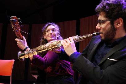 Auf dem Bild sind zwei musizierende Personen zu sehen. Die vordere Person spielt Saxophon und ist von der Seite abgebildet. Die hintere Person mit langen Haaren und einem roten Pullover spielt Gitarre und schaut fröhlich. 