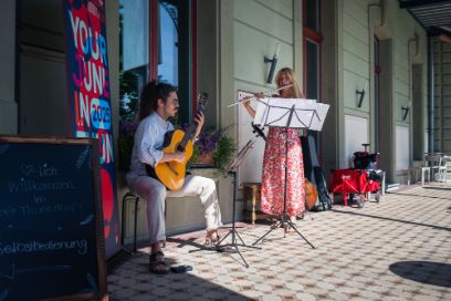Vor einem alten Gebäude auf einer Veranda mit schönem Plattenboden stehen zwei junge Personen, die musizieren. Die Person links spielt Gitarre. Die Person rechts trägt ein rotes Sommerkleid und spielt Querflöte. 