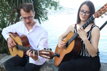 Vor einem Baum und einem See sitzen zwei junge Personen, die Gitarre spielen. 