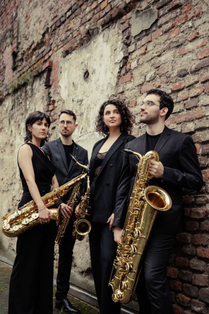 Vier Menschen vor einer Ziegelmauer, die etwas verfallen ist. Alle sind elegant gekleidet und tragen Saxophone in den Händen. 
