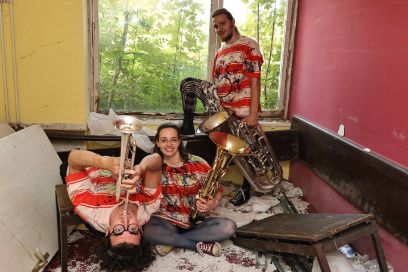 Drei Menschen sind in einem bunten Zimmer in einem baufälligen Haus, um sie herum ist Bauschutt. Das Fenster steht offen und man sieht einen Wald. Die Personen tragen rot gestreifte Shirts und alle haben ein Musikinstrument in der Hand. 
