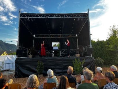 Konzertbühne mit drei Personen, einer Sängerin im roten langen Kleid, eine Frau an der Querflöte und einer dritten Person am Piano. 