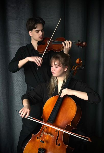 Zwei junge Menschen spielen ihr Instrument vor einem schwarzen Vorhang. Im Hintergrund ein Mann stehend mit Geige, im Vordergrund eine Frau sitzend mit Cello. Beide haben die Augen geschlossen.