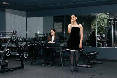 In einem Fitnessstudio zwischen Hanteln und einem Spiegel stehen zwei junge Frauen. Die vordere hält ein Mikrofon in der Hand und singt. Sie trägt ein elegantes, kurz geschnittenes schwarzes Kleid. Die hintere Frau sitz an einem E-Piano und schaut auf die Tasten. Sie trägt eine weisse Bluse und eine schwarze Jacke. 