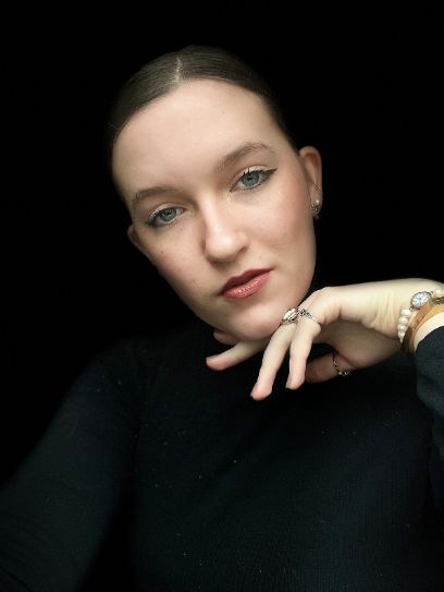 A young woman against a black background looks into the camera with her head slightly tilted, holding her left hand to her chin.