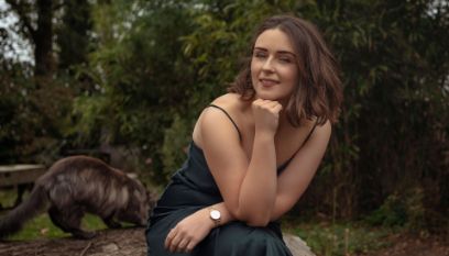 Fotoaufnahme einer sitzenden jungen Frau mit dunklem Kleid und dünnen Trägern. Ihr braunes Haar trägt sie offen. Das Gesicht ist abgestützt auf ihrer Faust. Am Handgelänk der anderen Hand ist eine Uhr zu sehen.