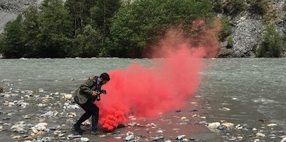 Fotograf am Flussbett in leicht geduckter Haltung, roter Rauch steigt direkt vor seinen Füssen auf.