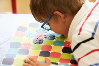 Kind mit Brille nah an Blatt mit bunten Vierecken, beschriftet mit Braille-Schrift