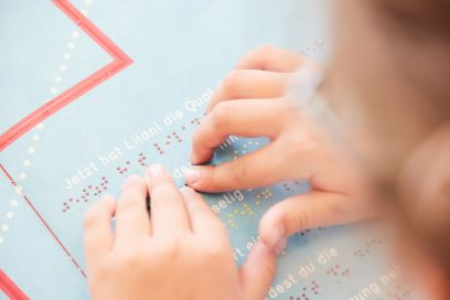 Kind mit Brille nah an Blatt mit bunten Vierecken, beschriftet mit Braille-Schrift
