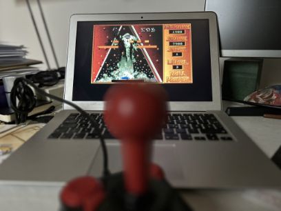 Abgebildet ist ein Joystick im Vordergrund und ein offener Laptop mit einer geöffneten Game-Applikation im Hintergrund.