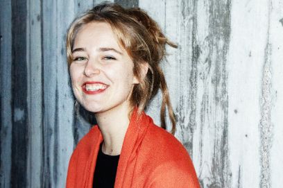 Portrait-Aufnahme der jungen Frau. Sie hat blondas, langes Haar, das sie zusammen gebunden trägt. Über einem schwarzen Oberteil trägt sie ein rotes, offenes Jäckchen. Sie lächelt mit offenem Mund in die Kamera.
