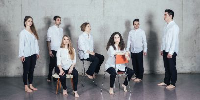 Sieben Musiker*innen des Ensembles, drei auf verschiedenen Stühlen sitzend, vier stehend, alle mit schwarzen Hosen und weissem Hemd und ohne Schuhe in einem grauen Raum mit Betonwand fotografiert.