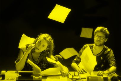An einem Bürotisch sitzen links eine Frau, rechts ein Mann. Auf dem Tisch sind Bürogeräte zu sehen. Papierblätter fliegen durch die Luft.