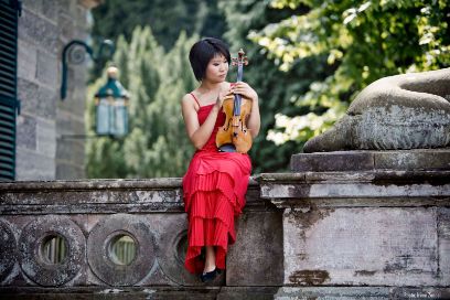 Tianwa Yang sitz in einem roten Kleid im Freien auf der steinernen Mauer eines Schlosses und hat ihre Violine aufgestützt.