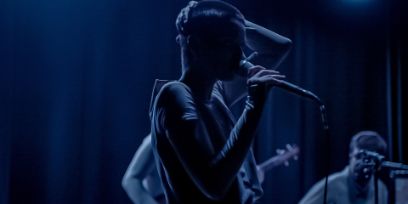 Prise de vue sombre et bleue. La jeune femme aux cheveux rasés se tient au centre de la photo, se tenant le crâne de la main gauche et portant le microphone à sa bouche de la main droite. En arrière-plan, on peut voir deux autres musicien-ne-s.