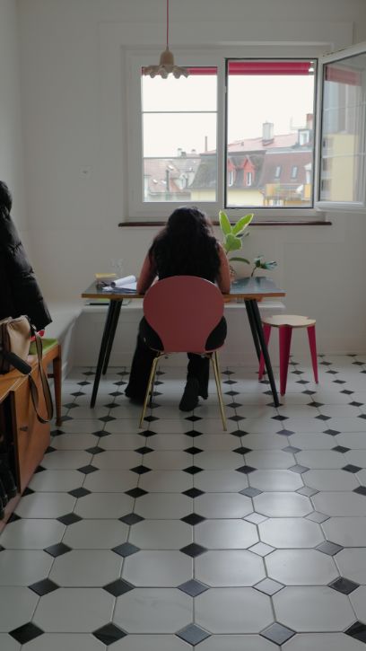 Frau sitzt an einem Tisch in Wohnung, Fliesenboden, lichtgefluteter Raum, Pflanzen