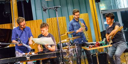 Vier Männer, spielen  Saxophon, E-Gitarre und Perkussion, sie sind im Gespräch