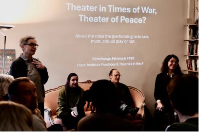 Aufnahme während des Vortrags. Menschen sitzen auf Stühlen und einem Sofa, im Hintergrund ist «Theater in Times of War, Theater of Peace?» an die Wand projiziert.