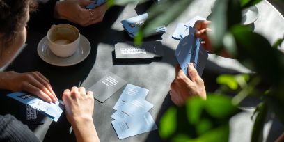 Drei Personen sitzen an einem Tisch, wir sehen die nur die Hände aus einer schrägen Vogelperspektive. Alle drei halten Karten in der Hand und spielen ein Spiel. Zudem ist eine Kaffeetasse zu sehen. 