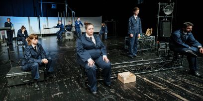 10 Personen sitzen oder stehen auf einer Bühne. Alle tragen dunkelblaue Overalls und haben eine andere Haltung.