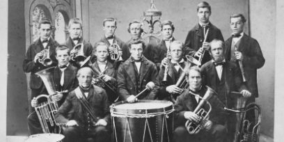 Eine schwarz-weiss Fotografie. 14 Männer schauen in die Kamera. Dabei halten Sie Blasinstrumente. Der Mann vorne in der Mitte hat eine grosse Trommel vor sich.