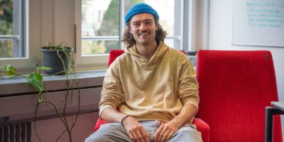 Le jeune homme a les cheveux bruns qui lui tombent jusqu'au menton, il porte un bonnet bleu de pêcheur et un sweat à capuche jaune beige. Il est assis sur un fauteuil de cinéma rouge devant une fenêtre. Il y a une plante sur le rebord de la fenêtre.