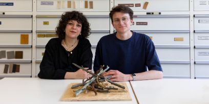 Zwei junge Personen sitzen vor einer weissen Wand mit vielen Schubladen. Links: Eine junge Frau mit lockigen, dunklen Haaren. Rechts: Ein junger Mann mit Brille. Im Vordergrund ist ein Teil ihrer Installation bestehend aus dünnen Ästen in verschiedenen Erdtönen aus Keramik.