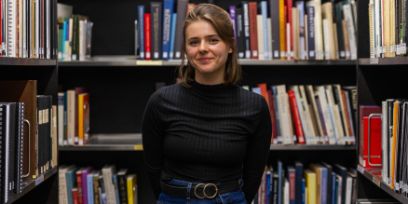 Eine Frau steht in einer Bibliothek. Links, rechts und hinten sind Bücher im Regal. Die Frau trägt ein schwarzes Oberteil und lächelt in die Kamera.