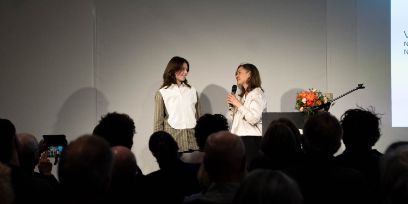 Zwei Frauen stehen vor einem Publikum. Vom Publikum sind die Hinterköpfe in dunklem Licht zu sehen. Violetty Dyka steht links, sie trägt eine Bluse in den Farben weiss und oliv. Dr. Minou Afzali steht rechts im Bild und hält ein Mikrofon – sie spricht.