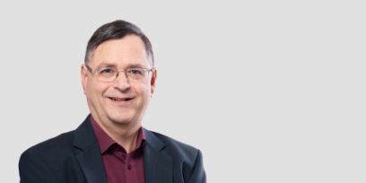 Ein Portrait eines Mannes mit Brille. Der Mann trägt ein bordeauxrotes Hemd und ein Jacket. Er lächelt.