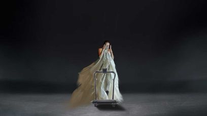 Eine Person in einem weissen Kleid steht auf einer dunklen Bühne und hält sich die Hände vors Gesicht. Sie steht dabei auf einem Laufband.
