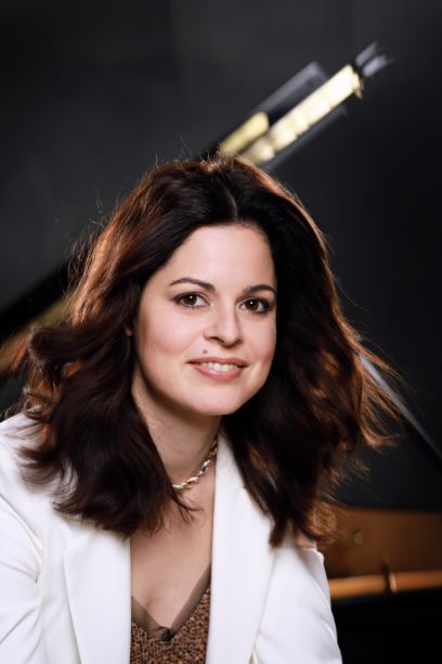 Eine Frau schaut direkt in die Kamera. Sie hat braunes Haar und ein weisses Jacket an. Sie lächelt.