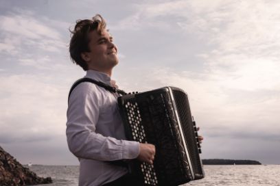 Ein Mann steht mit einem Akkordeon in der Hand an einem grossen Gewässer