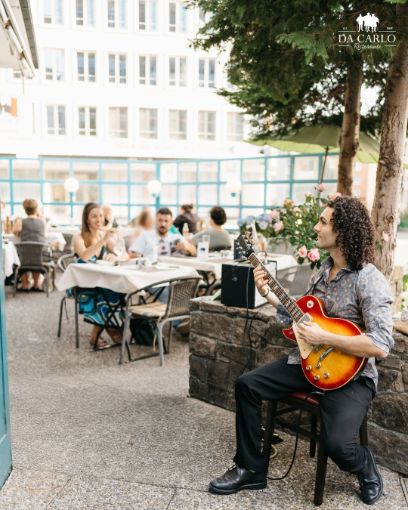 Rechts sitzt ein junger Mann (Marco Di Bari) in einem Restaurant und spielt auf einer elektrischen Gitarre. Im Hintergrund sitzen Menschen und hören zu.