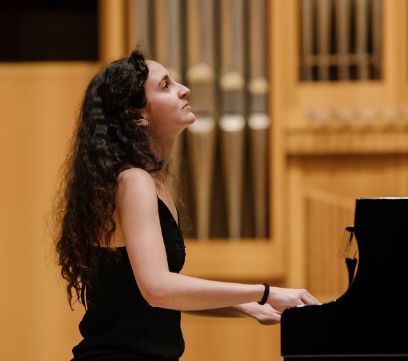 Eine Frau sitzt am Klavier. Sie hat dunkle Haare und ein ärmelloses schwarzes Kleid an. 
