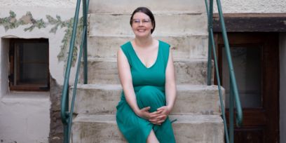 Die junge Frau sitzt auf der Treppe des Lenos Verlags. Ihr türkisfarbenes Kleid passt perfekt zum Geländer. Sie trägt das braune Haar nach hinten gebunden und eine Brille. 