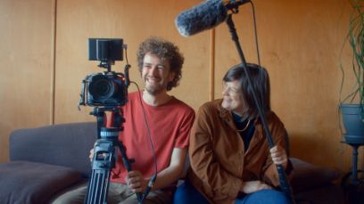 Luca Schmid und Bianca Schellander sitzen auf einem Sofa. Luca hält eine Kamera und Bianca ein Mikrofon, beide lachen.
