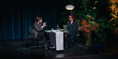 Zwei Frauen sitzen auf einer Bühne. Die Bühne ist mit Pflanzen und einem Licht dekoriert. 