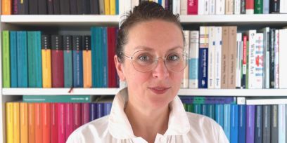 Portrait von Hanna B. Hölling. Sie ist in der Mitte des Bildes vor einem Bücherregal mit verschiedenfarbigen Büchern drin. Sie trägt die braunen Haare nach hinten gebunden, eine Brille und eine weisse Bluse.