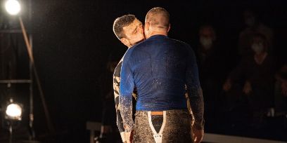 Bühnenszene: zwei Männer stehend und sich küssend. Der eine mit dem Rücken zur Kamera und der andere ihm gegenüber mit etwas zur Seite geneigtem Kopf und geschlossenen Augen.