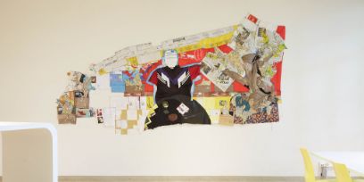 Verschiedene Gegenstände, wie Kartonverpackungen, Zeitungen, alte Kleider, sind an einer weissen Wand angeordnet und ergebem ein Bild von einer Person die an einem Tisch sitzt.