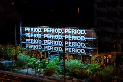 Aufnahme der Installation bei Nacht. Das Wort «PERIOD.» steht jeweils dreimal hintereinander in vier Zeilen in leuchtendem Hellblau. Darunter erkennt man grünes Gebüsch und Bäume, dahinter ein Gebäude in warmen Orange- und Gelbtönen.