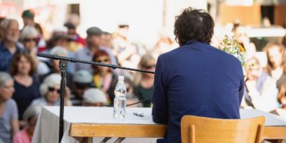 Schriftsteller sitzt draussen an einem Tisch, eine Wasserflasche und ein Stift ist zu seiner Linken zu sehen. Neben dm Tisch steht ein Mikrophon-Ständer. Der Schriftsteller ist von hinten abgelichtet und vor ihm, im Hintergrund der Aufnahme sitzt ein unscharf aufgenommenes Publikum